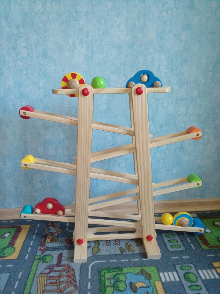 Eine hölzerne Murmelbahn, auf der außer Murmeln auch zwei Holzautos herunterfahren. Auf dem Boden liegt ein Teppich, auf dem Straßen, Häuser und Parks aufgedruckt sind