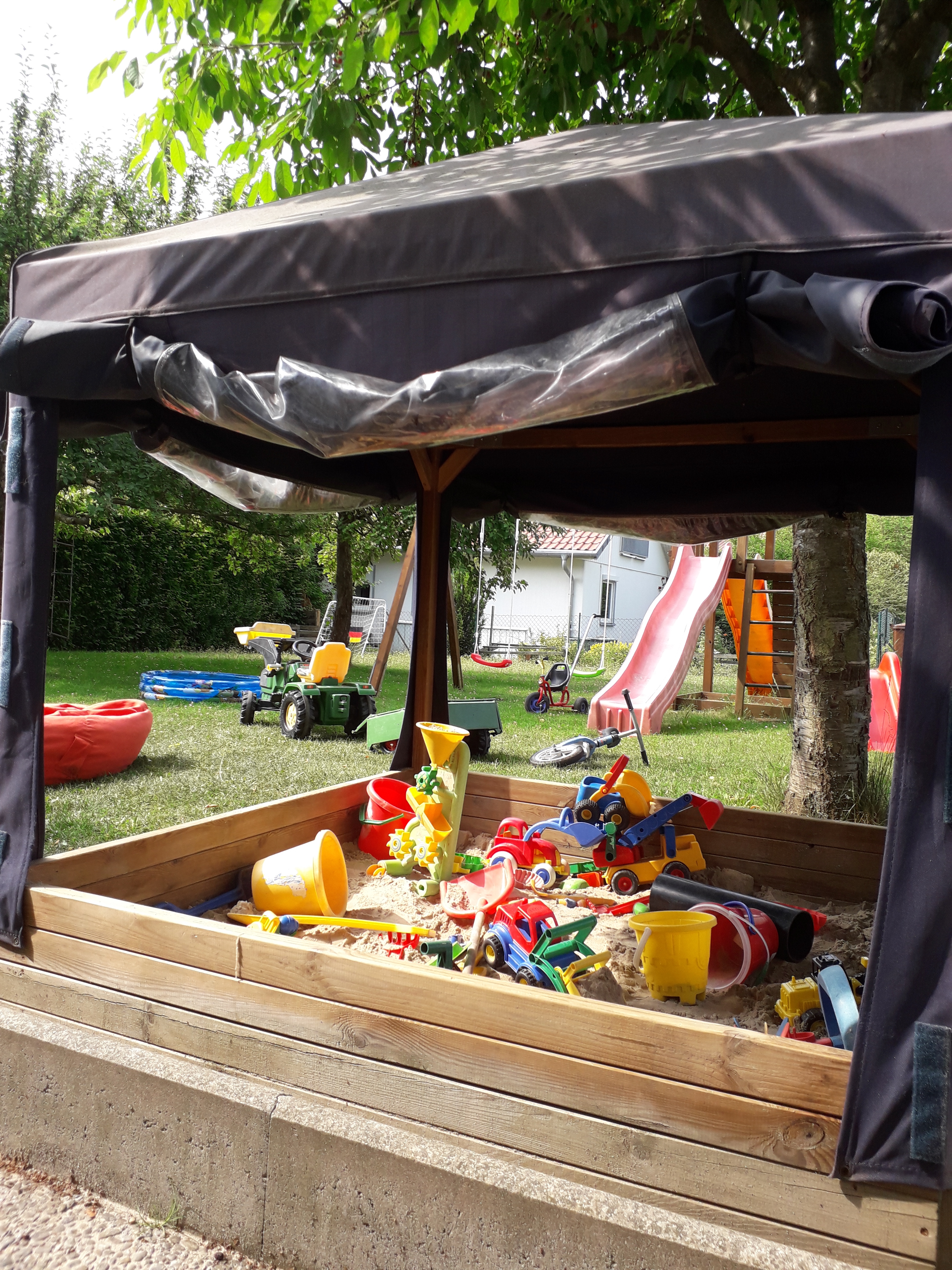 Ein Blick in unseren Garten - im Vordergrund ist ein großer Sandkasten mit Pavillion darüber und sehr vielen bunten Sandspielsachen darin, im Hintergrund sind ein Sitzsack, ein Plantschbacken, ein Klettergerüst mit Schaukeln und Rutschen und ein Laufrad zu sehen.