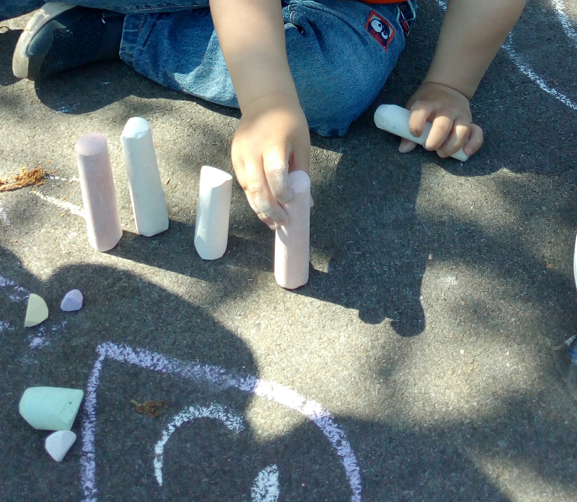 Eine Kinderhand, die Straßenmalkreiden nebeneinander aufstellt, auf dem Boden darunter ist ein mit diesen Kreiden gemaltes Herz zu sehen